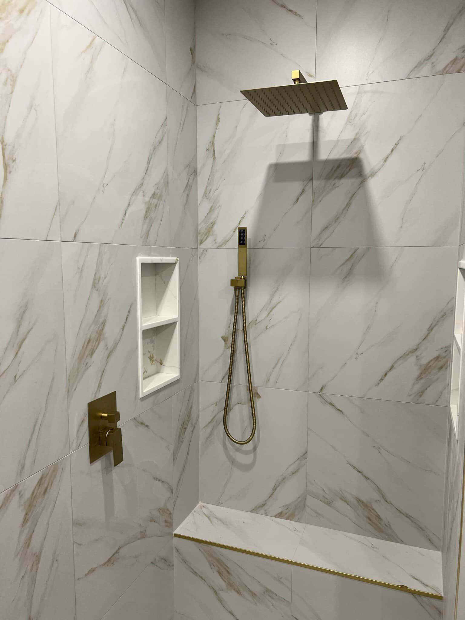 Une douche récemment carrelée avec des accessoires dorés. Carrelage imitation marbre blanc veiné d'or, pommeau de douche et étagère intégrée.