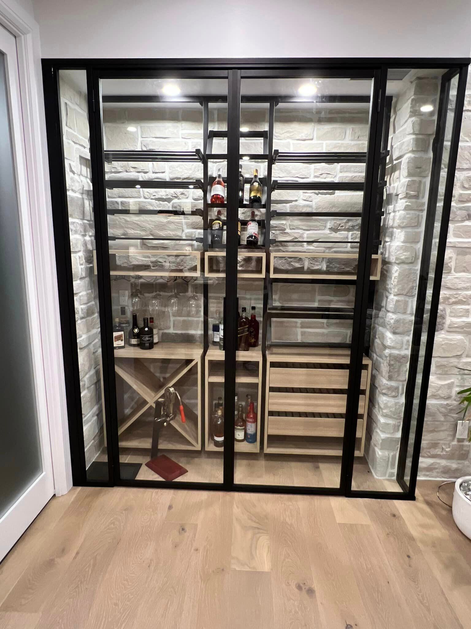 Cave à vin avec porte vitrée et cadre en métal noir. Les bouteilles sont exposées sur des étagères contre un mur de briques.