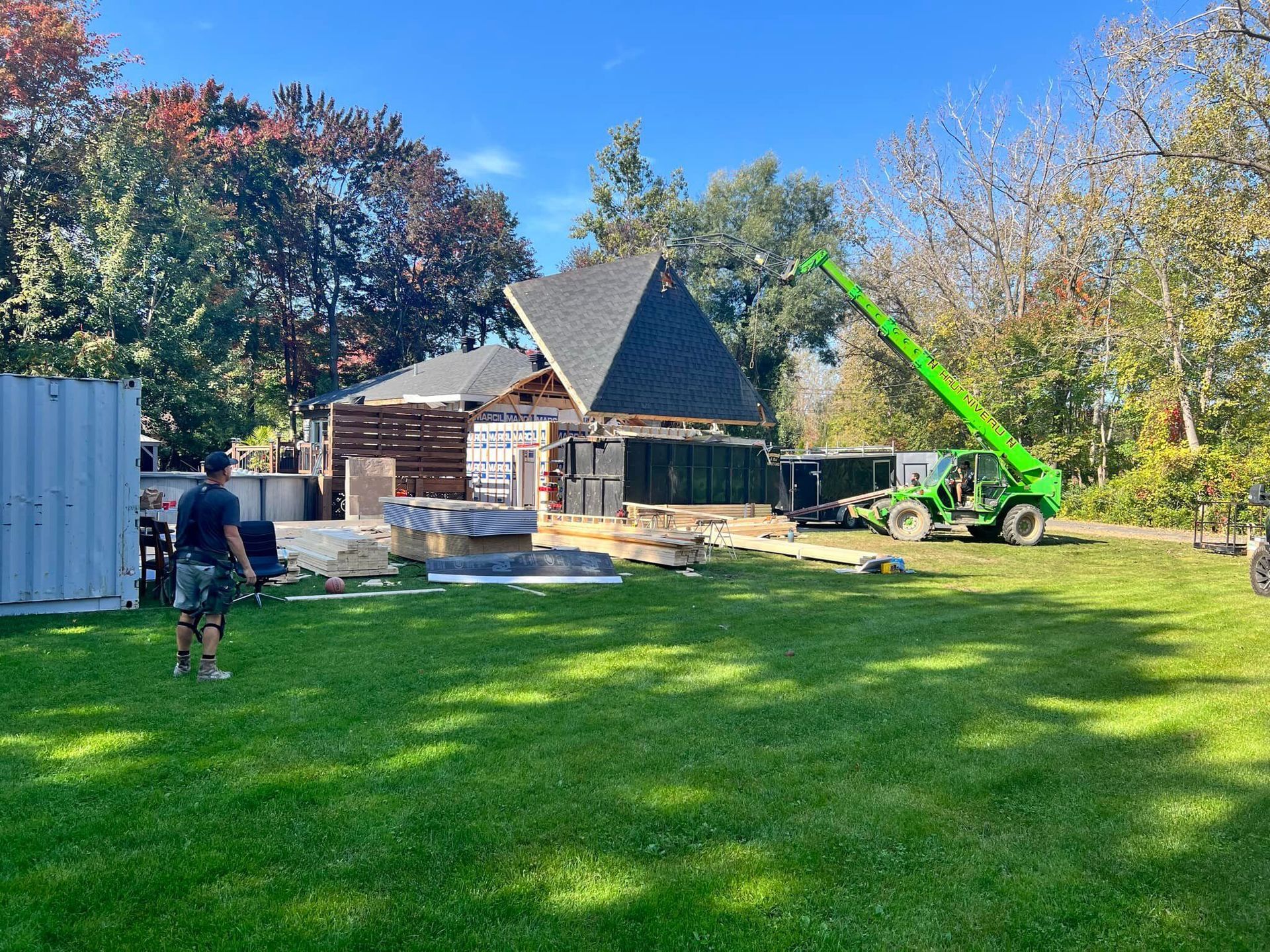Chantier de construction avec un bâtiment partiellement construit, un chariot élévateur vert et un ouvrier sur une zone herbeuse.