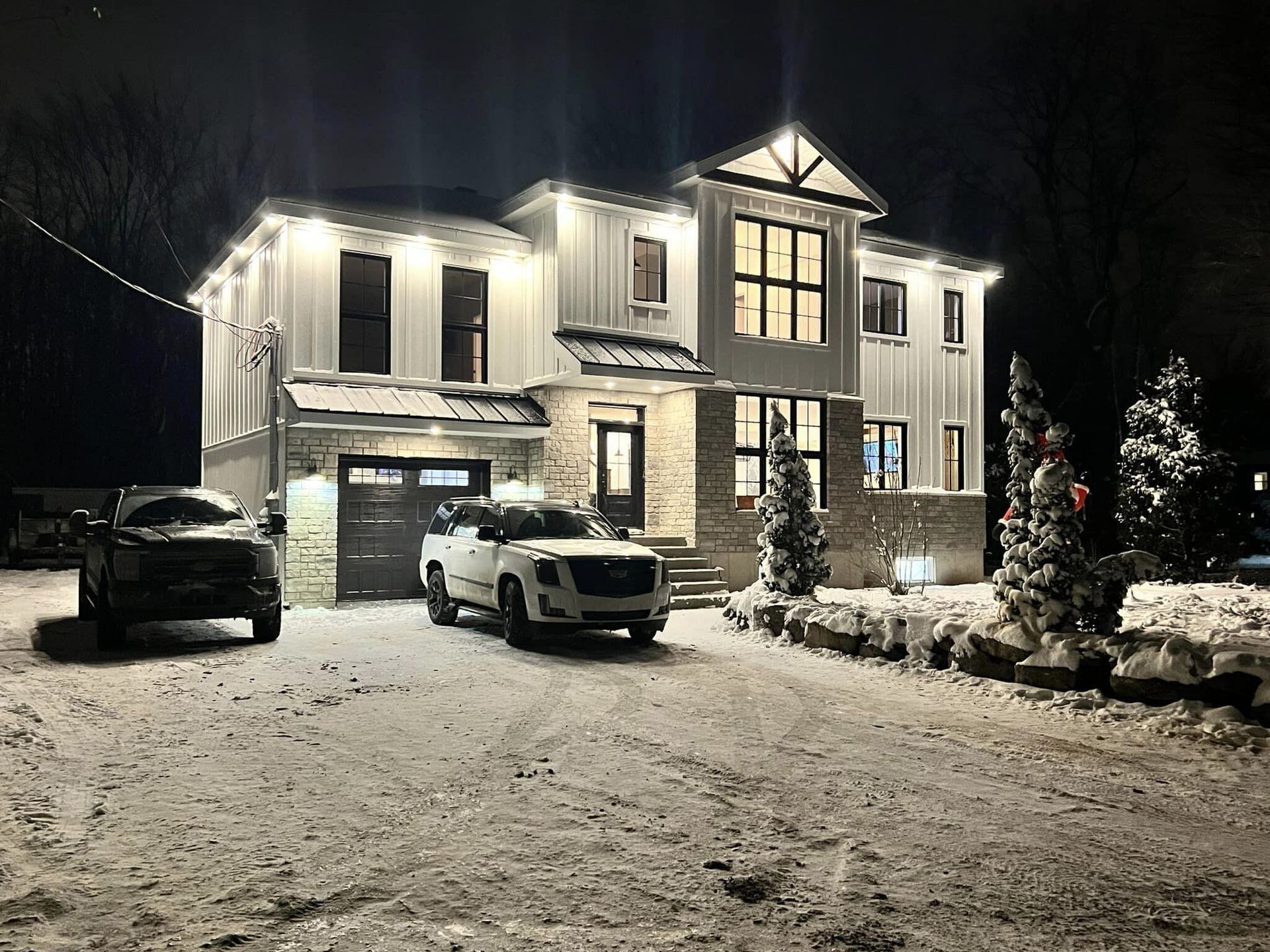 Une maison enneigée la nuit avec des lumières de Noël et deux camionnettes dans l'allée.