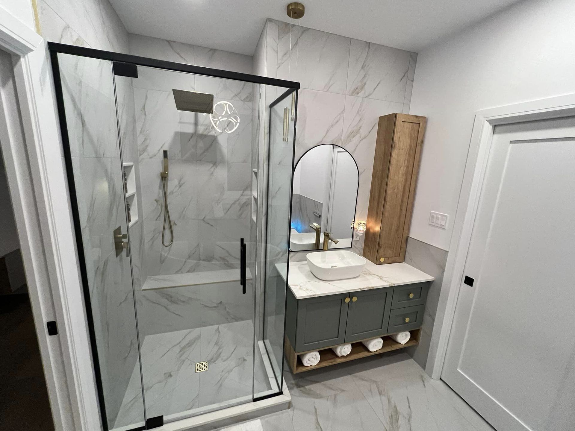 Salle de bain moderne avec douche en verre, meuble-lavabo et carrelage imitation marbre. Elle est également dotée d'une armoire en bois et d'un miroir bombé.