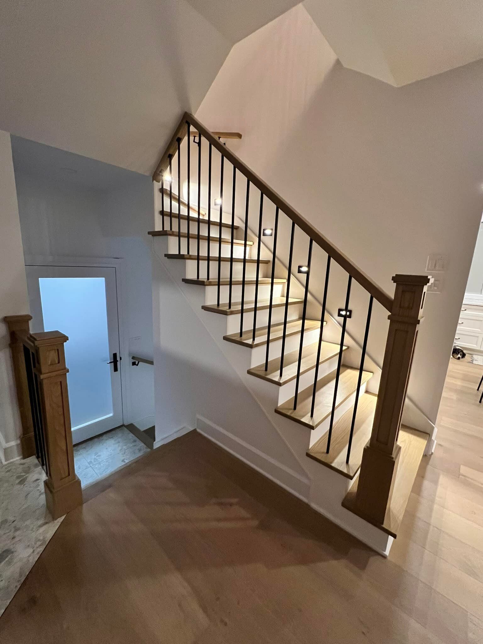 Vue intérieure d'un escalier en bois avec rampes noires et murs blancs, menant à une porte en bas.