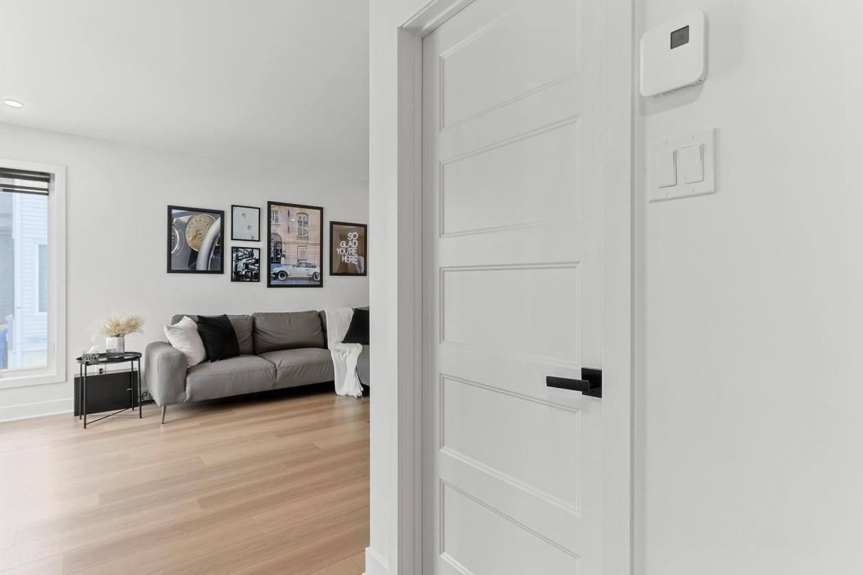 Porte blanche partiellement ouverte, menant à un salon avec un canapé gris, un mur de galerie et du parquet.