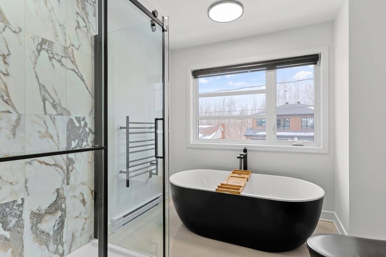 Salle de bain moderne avec baignoire noire, douche carrelée en marbre et fenêtre donnant sur un quartier.