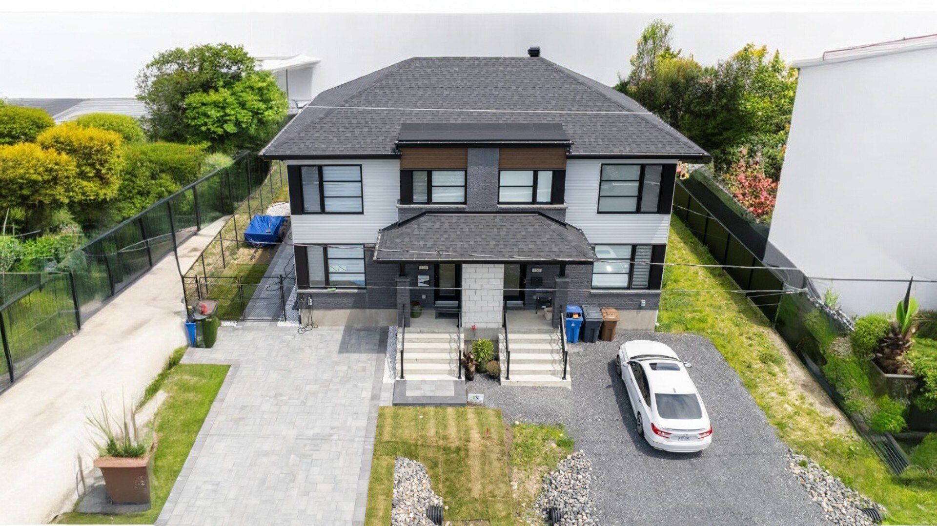 Duplex à deux étages avec toit gris foncé et façade gris clair. Allée avec voiture garée devant. Pelouse verte et verdure alentour.