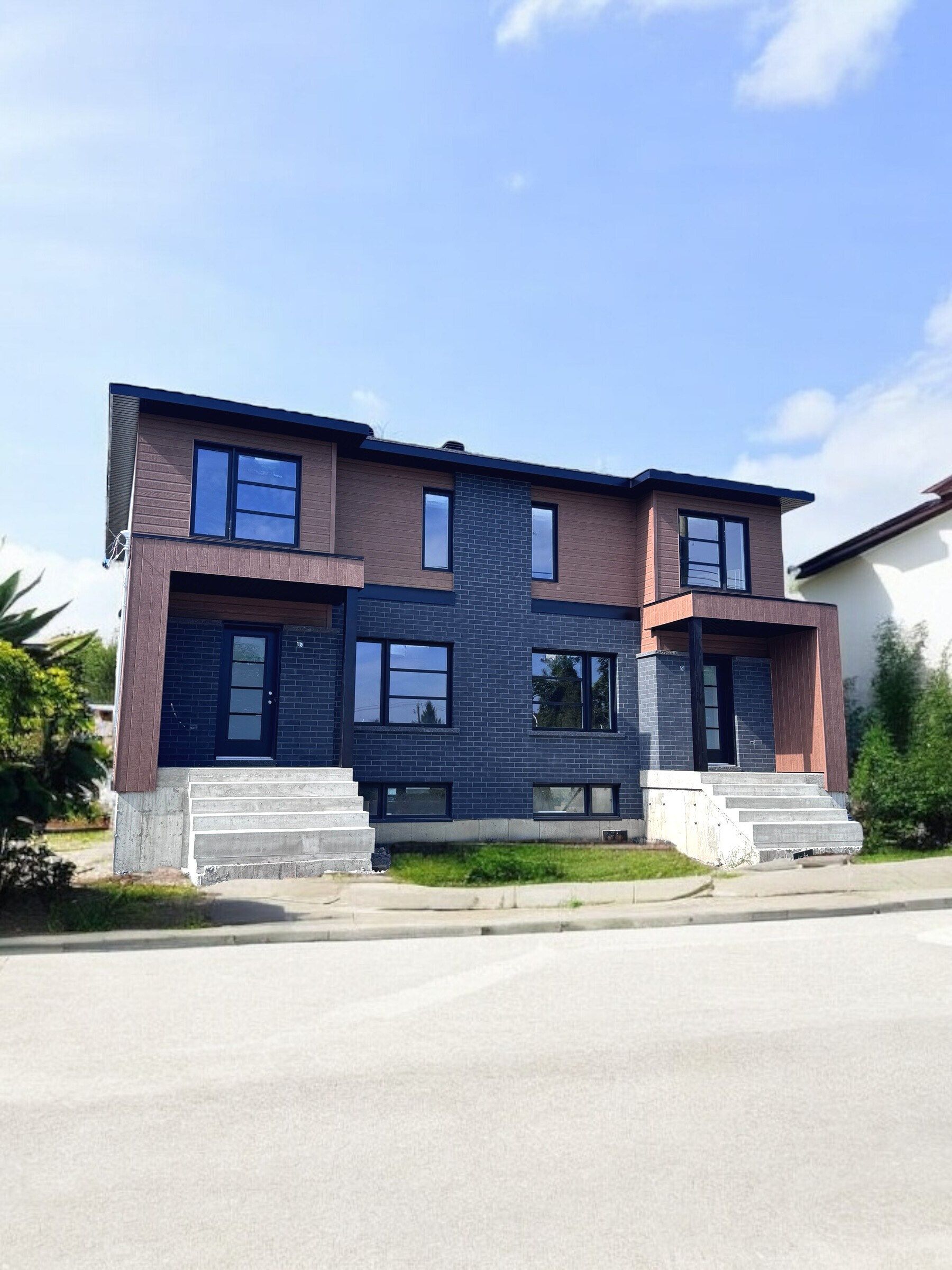 Duplex moderne de deux étages avec cadres de fenêtres sombres, façade grise et brune, posé sur une fondation en béton sous un ciel bleu.