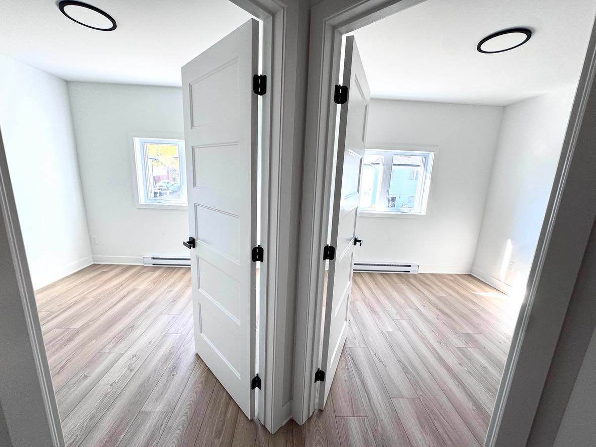 Deux portes blanches s'ouvrent sur des pièces identiques avec parquet, fenêtres et murs lumineux.