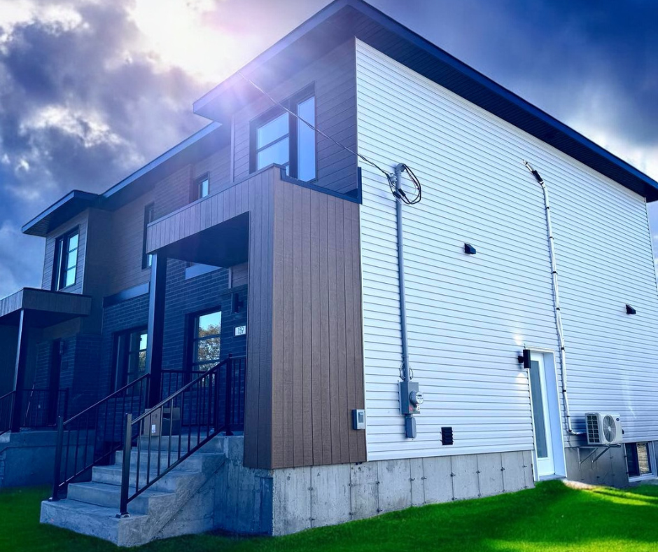Maison de ville moderne avec revêtement marron et blanc, touches de noir et un escalier menant à l'entrée. Le ciel bleu perce les nuages.