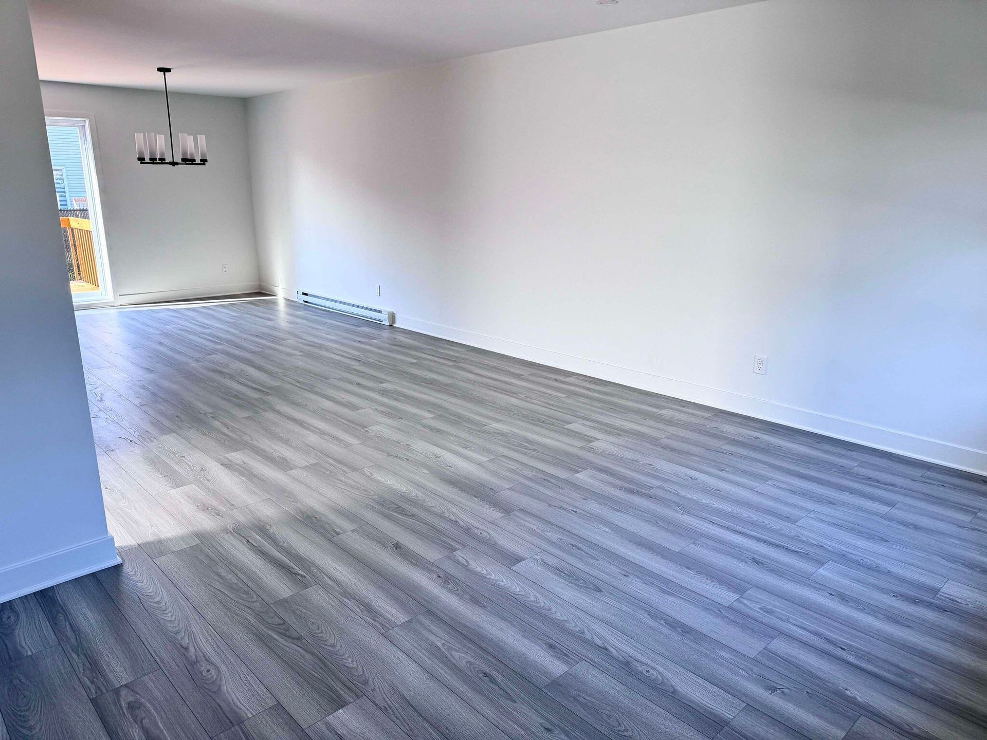 Pièce vide avec parquet gris, murs blancs et lustre noir moderne. Une porte mène à un espace extérieur.