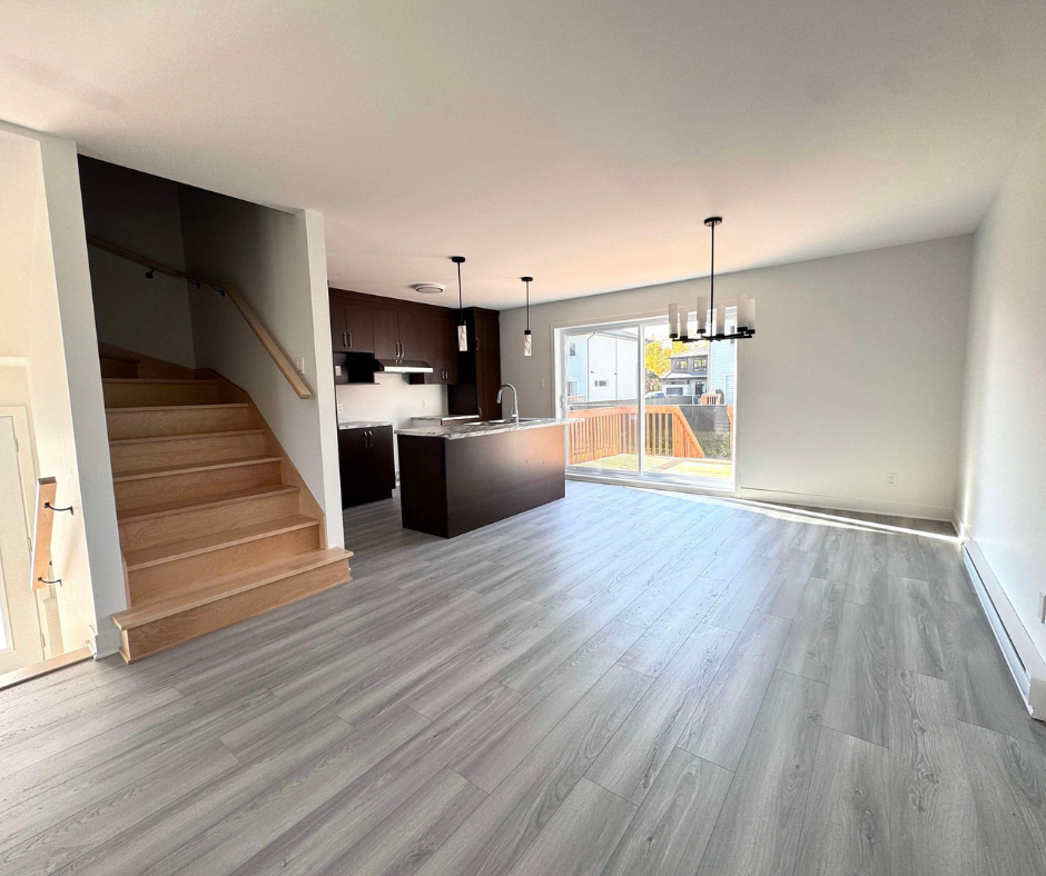 Intérieur à aire ouverte avec parquet imitation bois gris, armoires en bois foncé, escalier et porte coulissante donnant sur une terrasse ; les murs sont neutres.