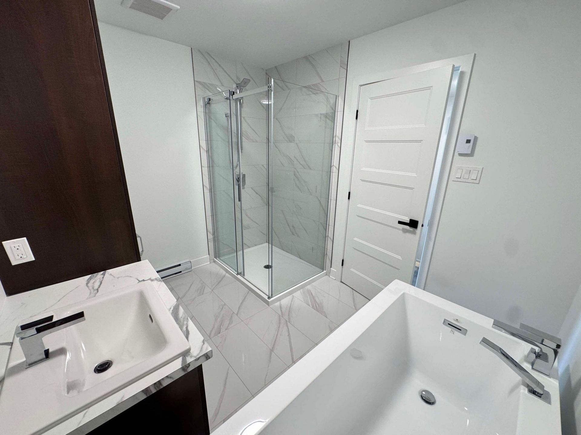 Salle de bain moderne avec baignoire blanche, douche en verre et sol imitation marbre. Un lavabo blanc se trouve à gauche et la porte à droite.