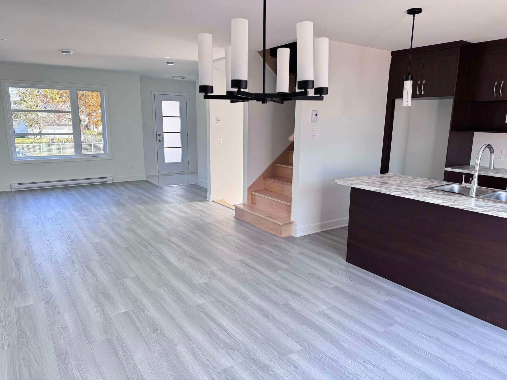 Un espace de vie ouvert avec un plancher en bois gris clair, une cuisine avec des armoires marron foncé et un lustre noir.