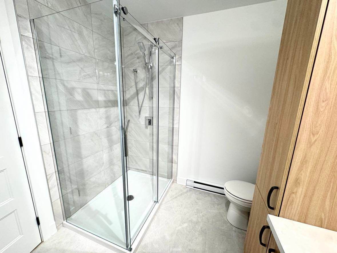 Salle de bain moderne avec douche en verre, toilettes et meuble en bois. Murs et sol en carrelage gris.