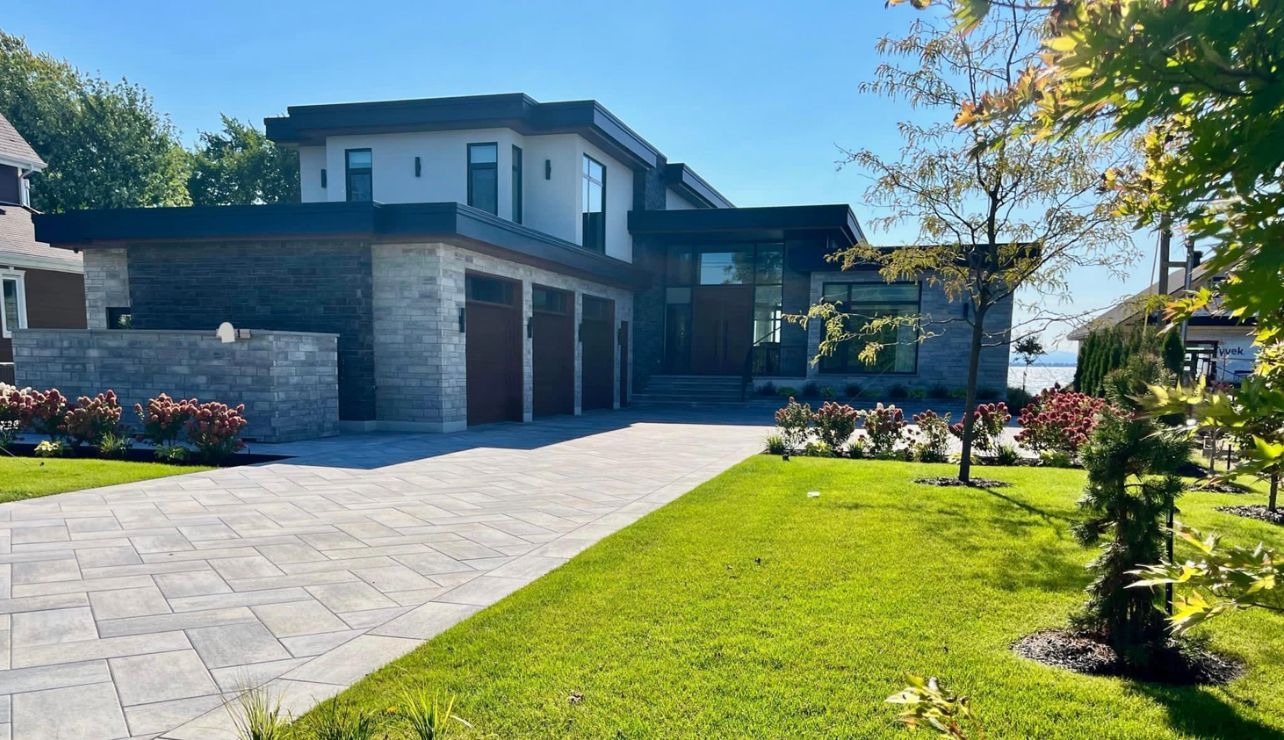 Maison moderne avec garage pour trois voitures et façade en pierre, sur une pelouse bien entretenue avec une allée pavée par une journée ensoleillée.