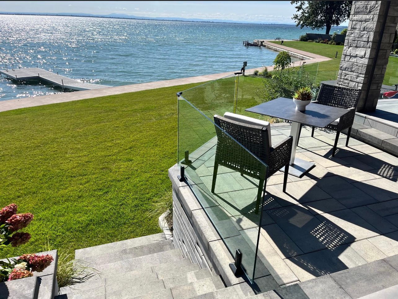 Terrasse avec table et chaises donnant sur un lac, avec balustrade en verre. Pelouse verdoyante et eau turquoise.