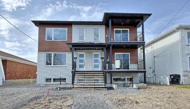 Duplex à deux étages avec revêtement gris et brun, deux entrées avec marches et balcons, situé sur un terrain en gravier.