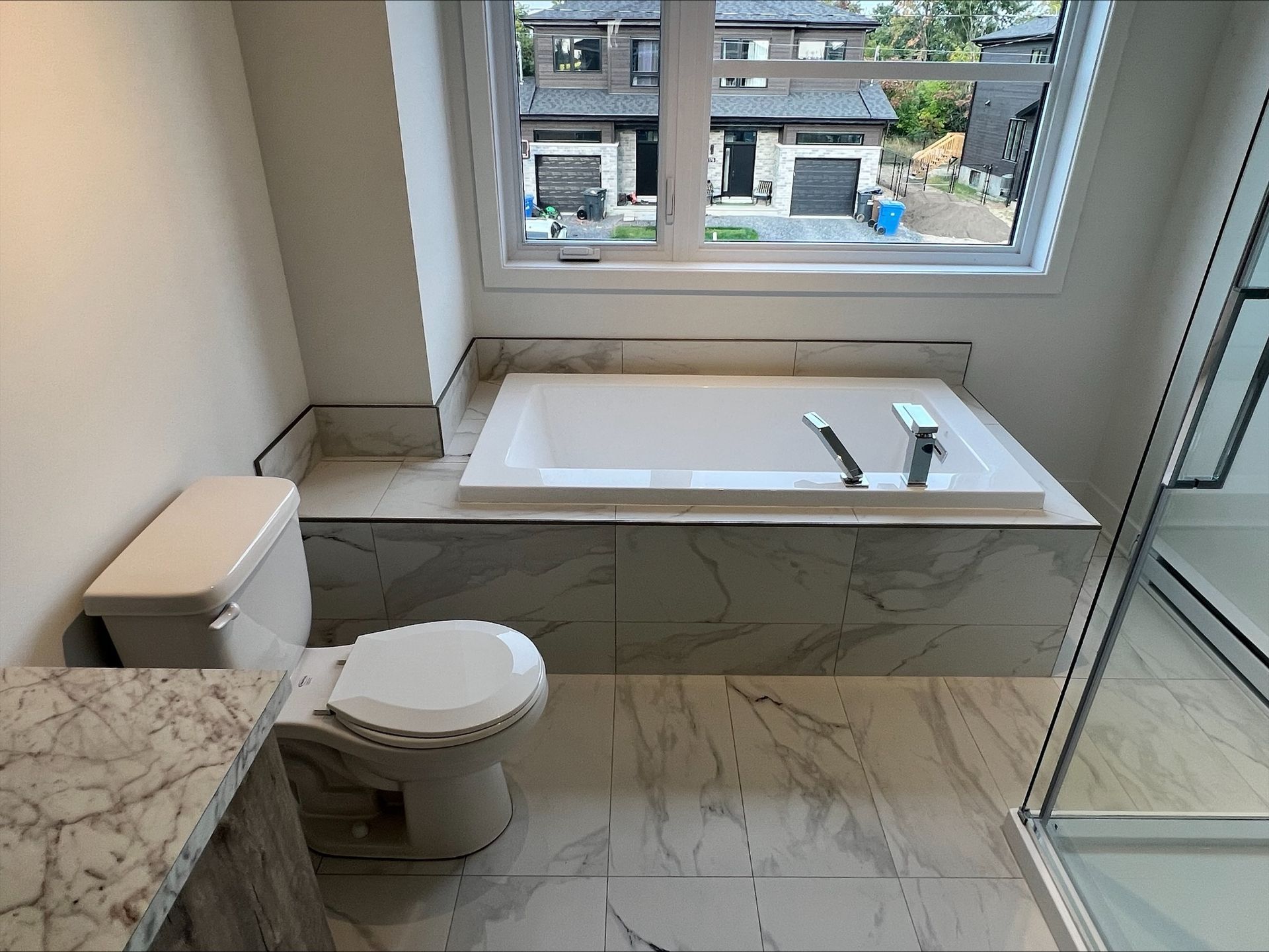 Une salle de bain avec baignoire blanche, toilettes et carrelage effet marbre. Une fenêtre offre une vue sur les maisons extérieures.