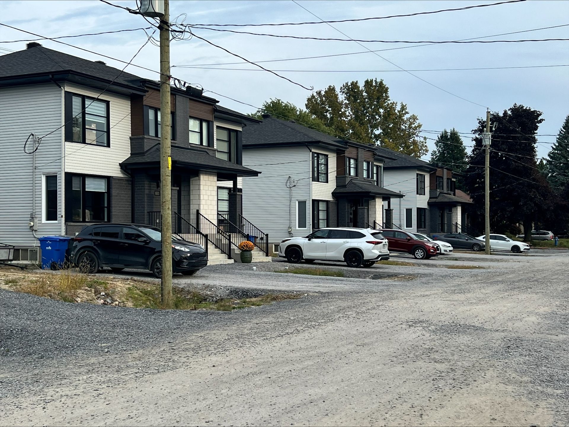 Rangée de maisons de ville modernes avec des voitures garées devant sur un terrain en gravier, sous un ciel couvert.