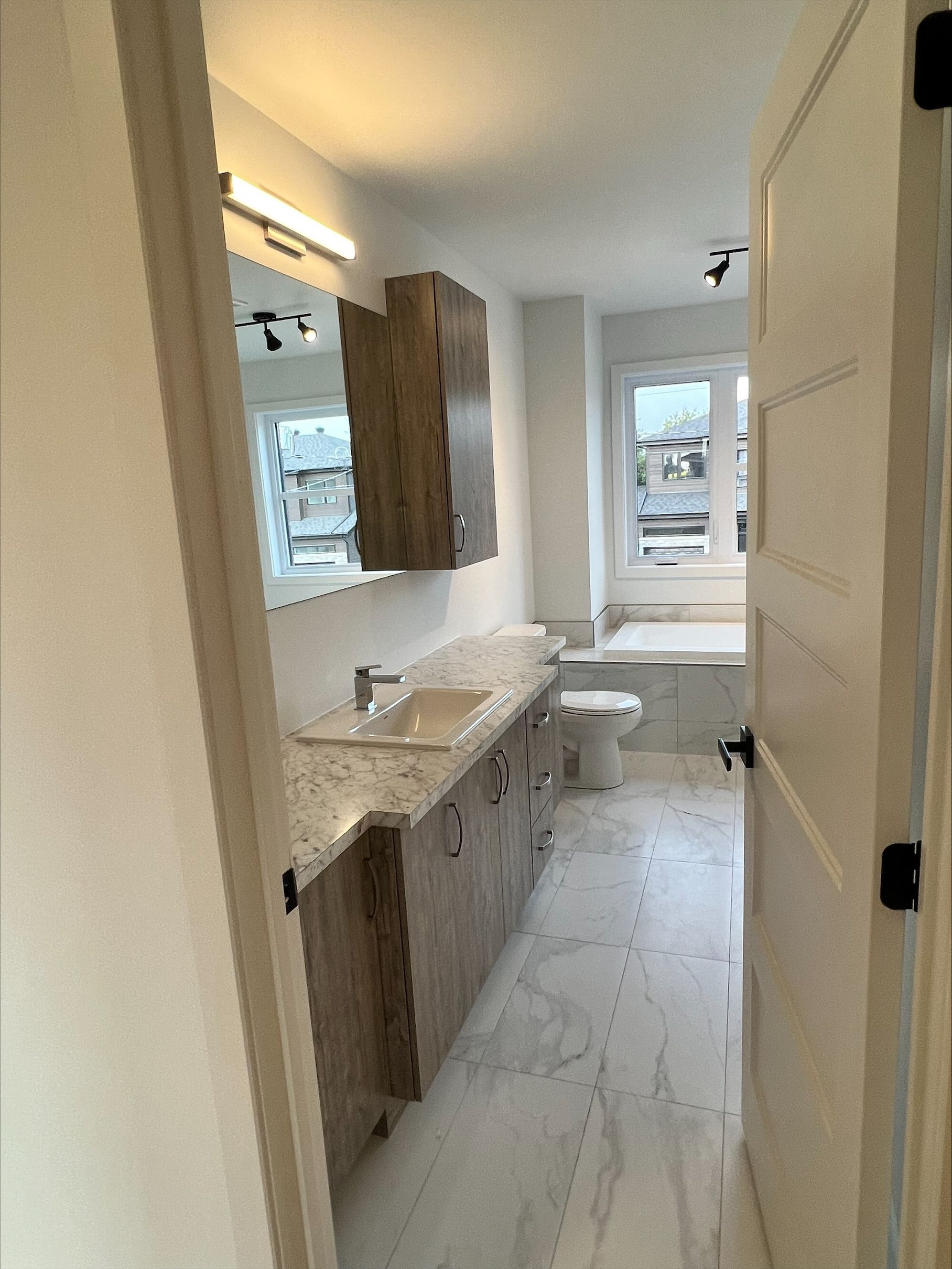 Salle de bains avec meubles clairs, sol effet marbre, fenêtre et toilettes. Vue depuis une porte.