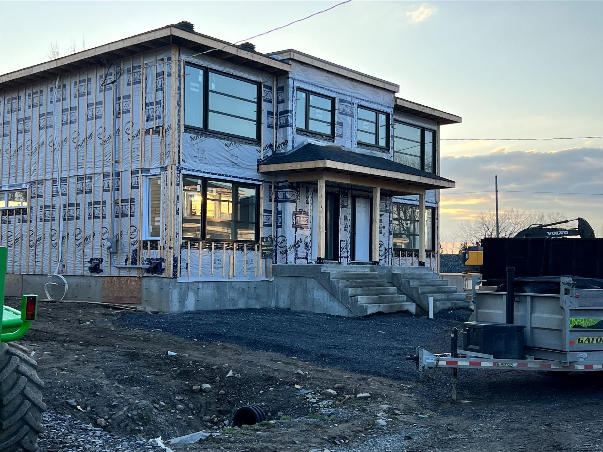 Une maison à deux étages en construction, avec charpente et revêtement en bois apparents, de grandes fenêtres et un petit porche avec marches.
