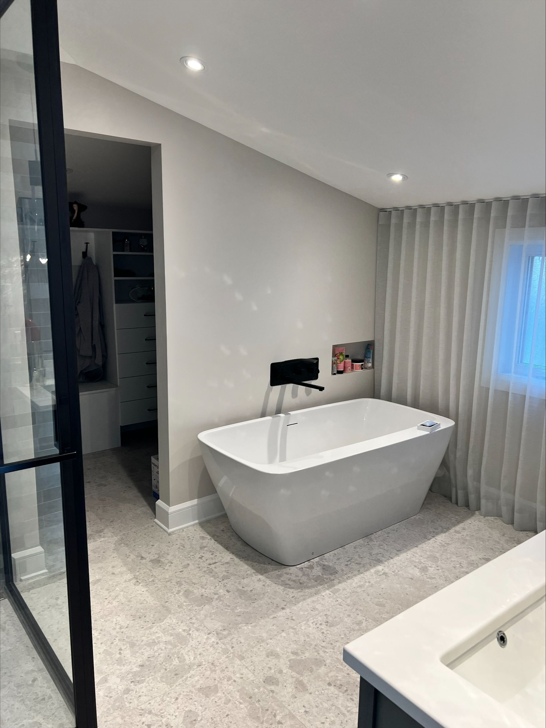 Salle de bain moderne avec une baignoire blanche autoportante, des murs gris et un dressing visible en arrière-plan.