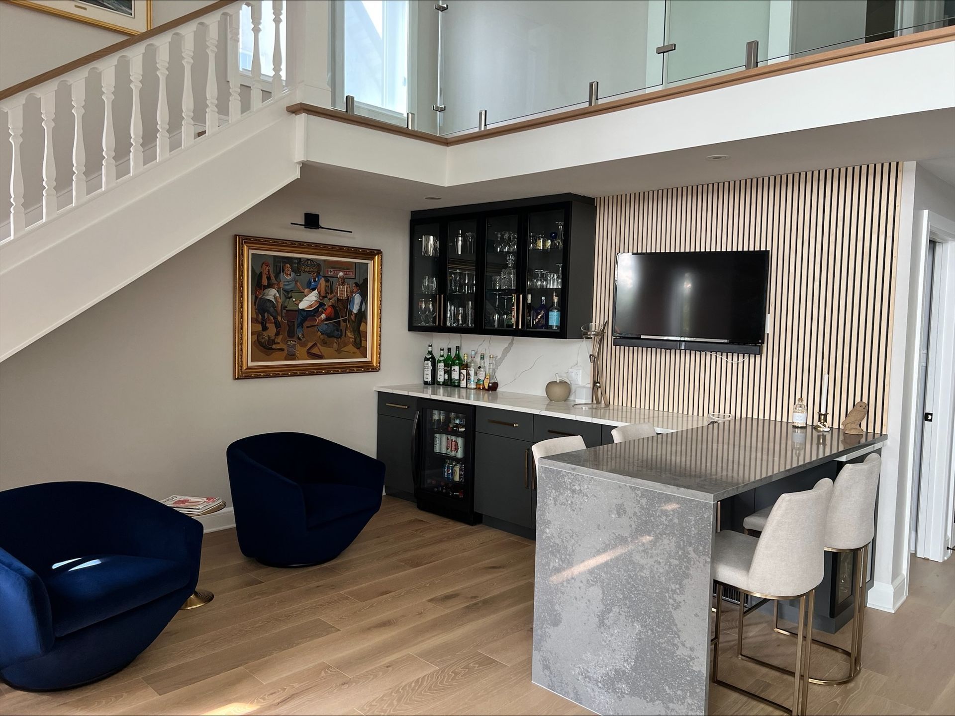 Un bar moderne avec un comptoir en pierre grise, des chaises, des meubles et une télévision. Deux fauteuils bleus sont installés près du bar, sous un escalier.