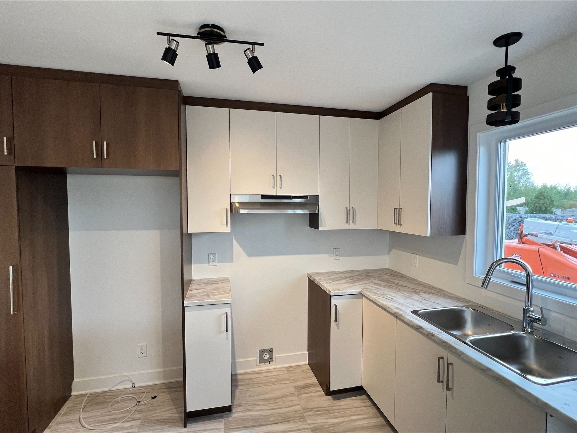 Cuisine avec armoires blanches et marron, évier en acier inoxydable et fenêtre. Luminaires apparents au plafond.