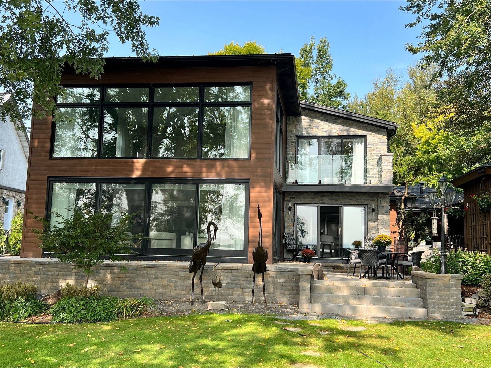 Maison moderne à deux étages avec de grandes fenêtres, revêtement en pierre et en bois, balcon et patio donnant sur une pelouse verte.