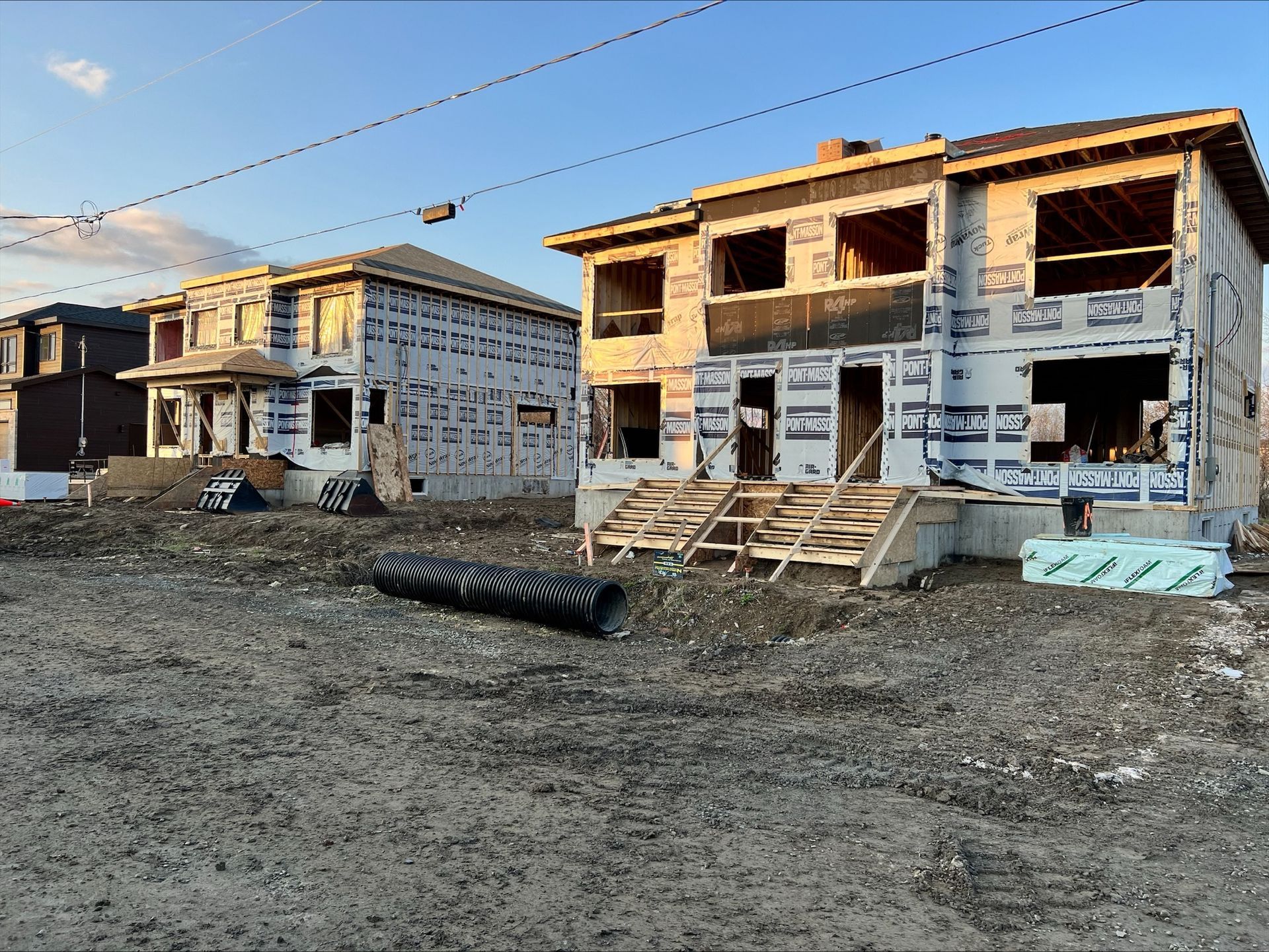 Deux maisons en construction, enveloppées d'un revêtement bleu, se trouvent sur un terrain en terre sous un ciel bleu.