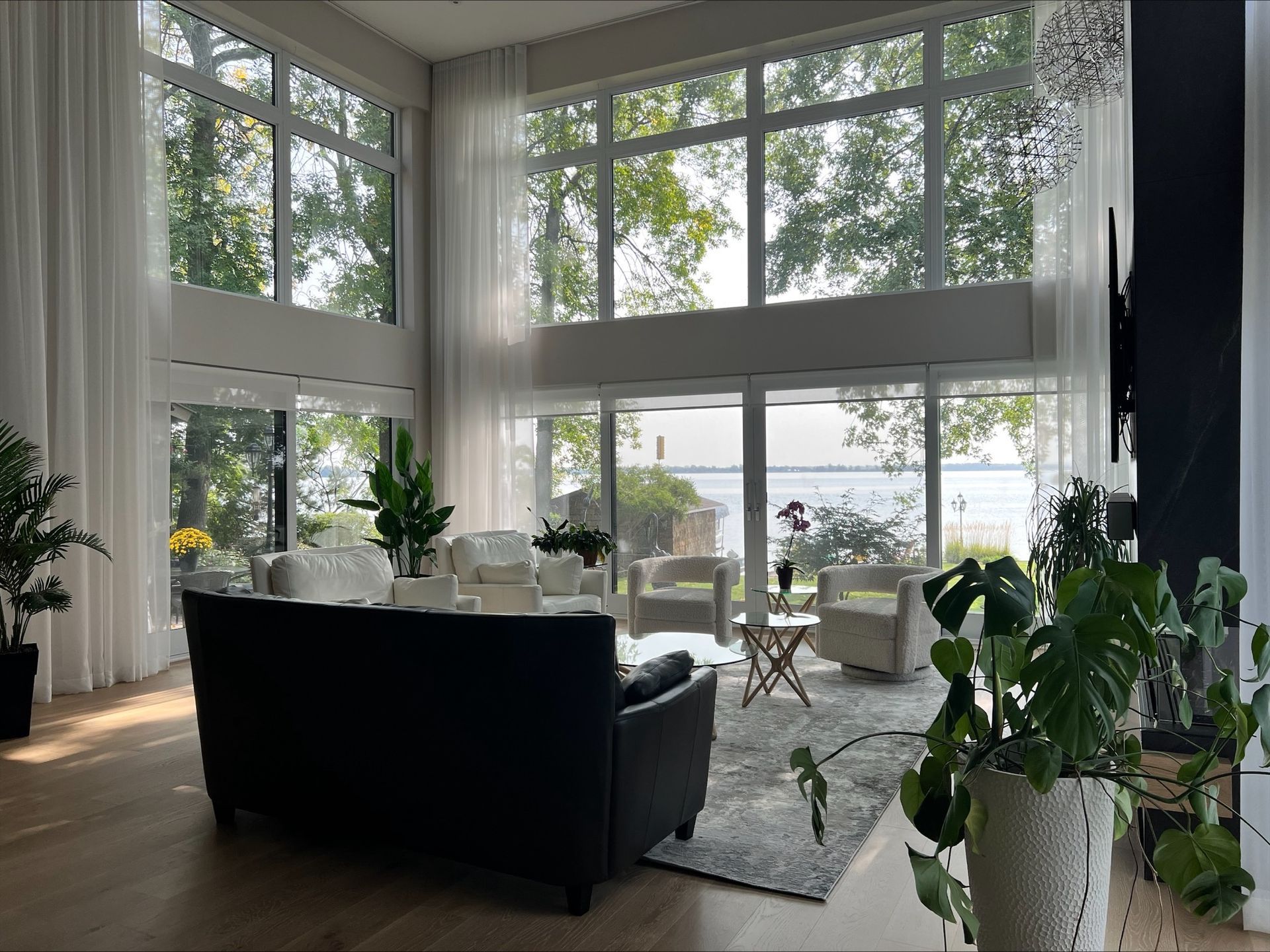 Salon lumineux avec baies vitrées donnant sur l'eau. Canapé noir, chaises blanches et plantes vertes complètent la pièce.