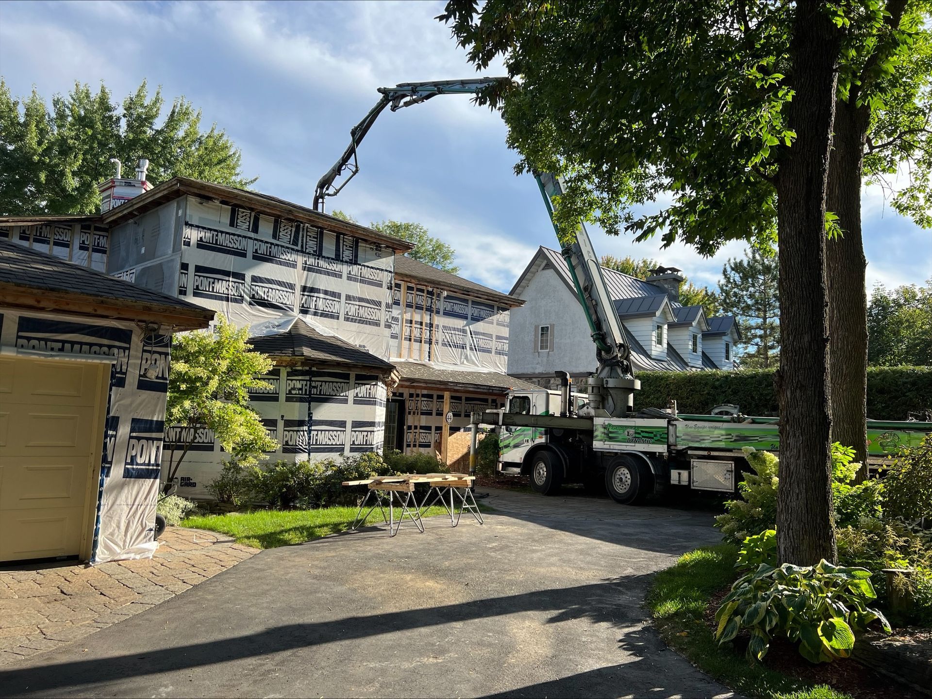 Un camion-pompe à béton coule du béton sur une maison en construction. La maison est isolée, avec des arbres et une pelouse au premier plan.