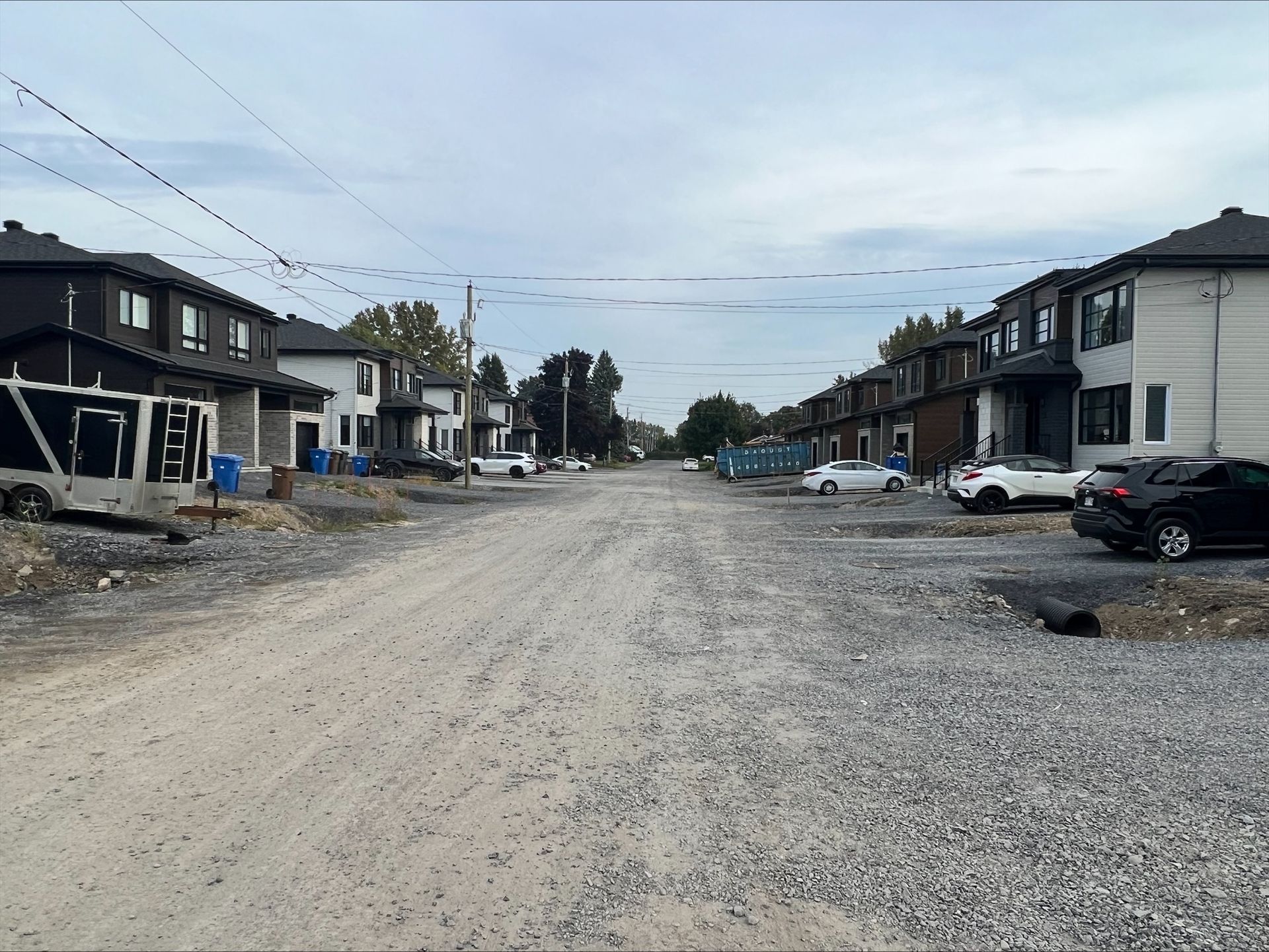 Une route de gravier dans un quartier résidentiel avec de nouvelles maisons de ville des deux côtés, des véhicules garés dans la rue et des lignes électriques aériennes.