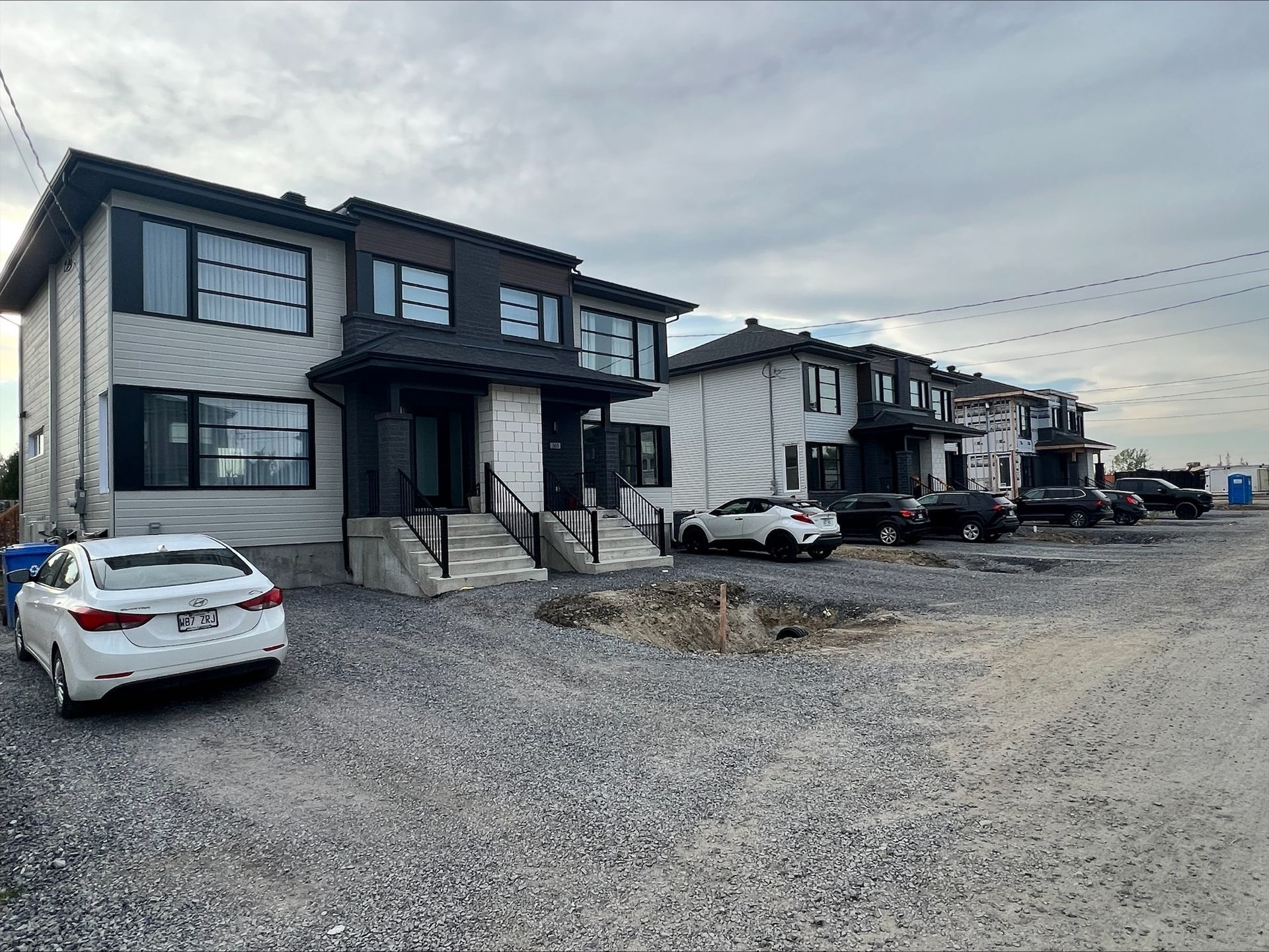 Maisons de ville modernes à deux étages aux touches sombres et aux grandes fenêtres sous un ciel nuageux. Allée de gravier avec voitures garées devant.