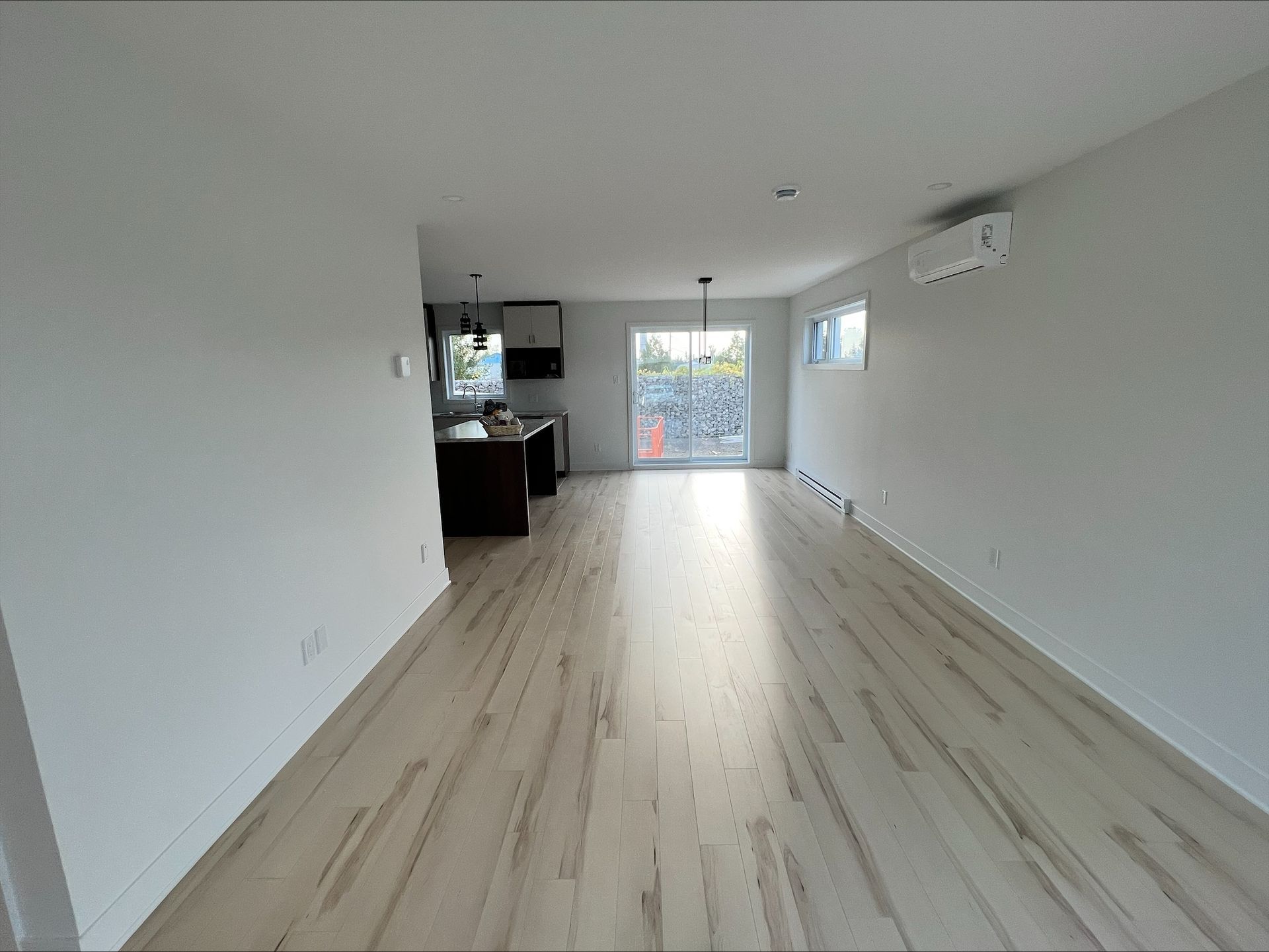 Intérieur d'un appartement vide avec parquet, murs blancs et une cuisine visible en arrière-plan. La lumière du soleil pénètre par une porte coulissante en verre.