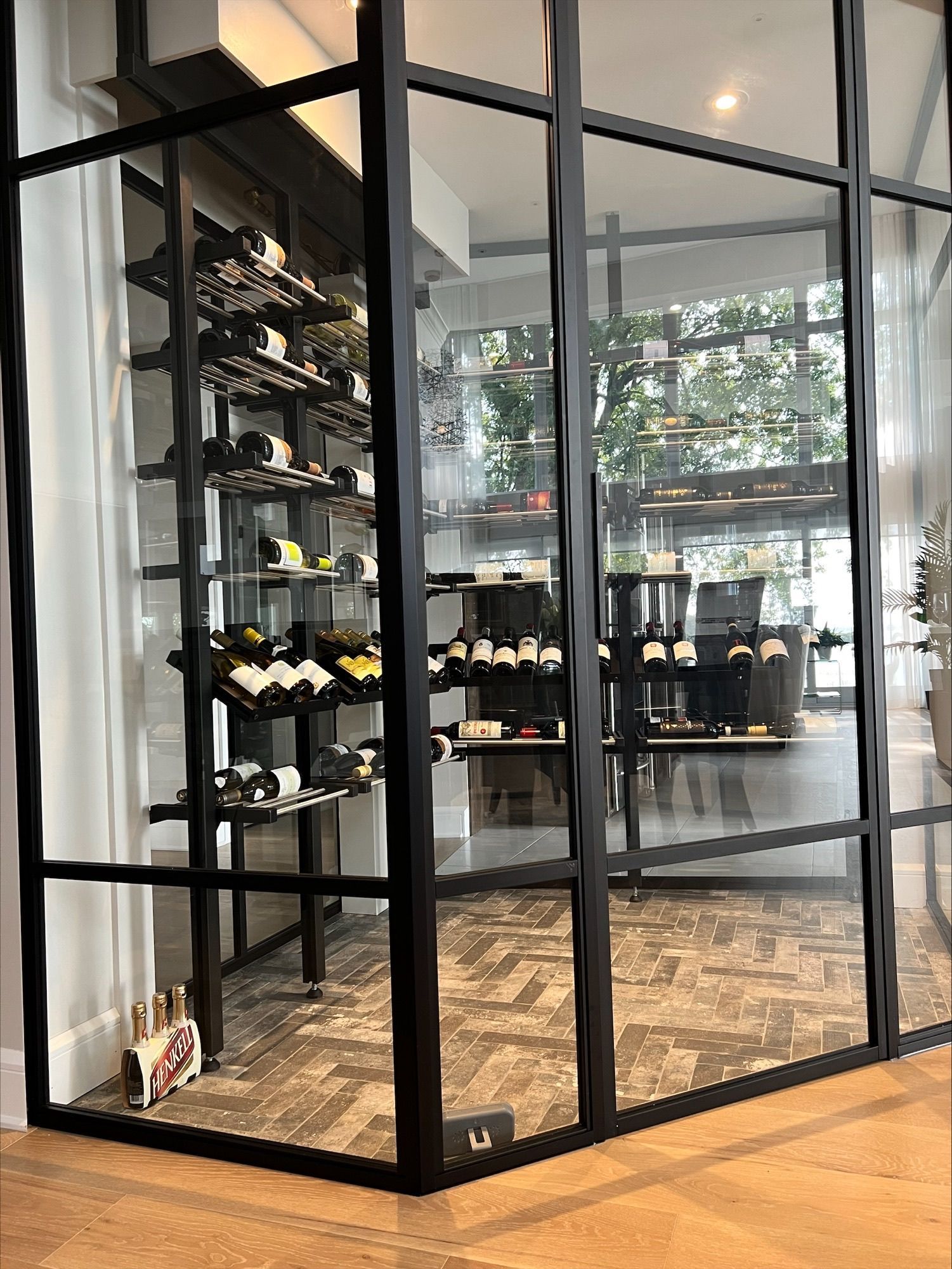 Une cave à vin aux parois vitrées aux cadres noirs. À l'intérieur, les bouteilles sont présentées sur des étagères. La pièce est dotée de parquet.
