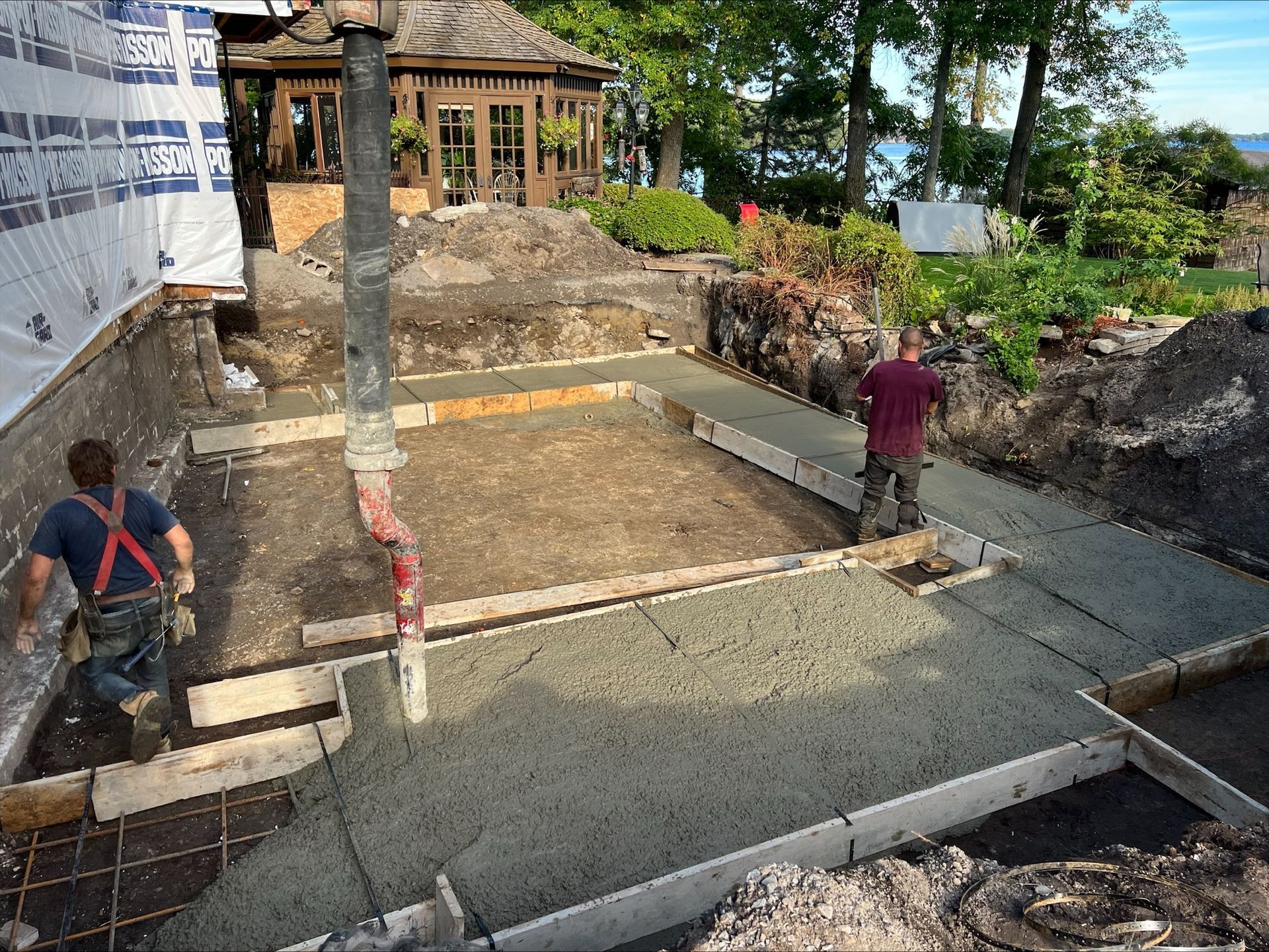 Des ouvriers du bâtiment coulent les fondations d'une structure, avec un belvédère et de l'eau en arrière-plan. Un ouvrier travaille sur le béton tandis qu'un autre s'éloigne.