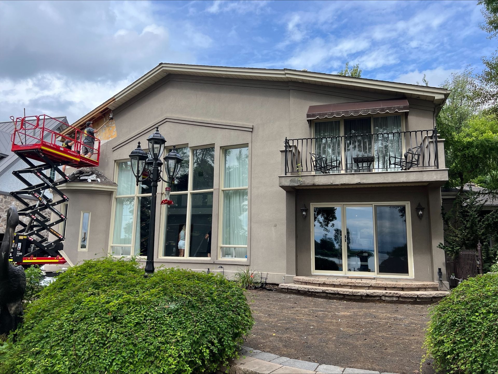 Une maison beige avec de grandes fenêtres dispose d'une plateforme élévatrice près du toit, avec une personne à l'intérieur. Un balcon se trouve au-dessus des fenêtres inférieures.