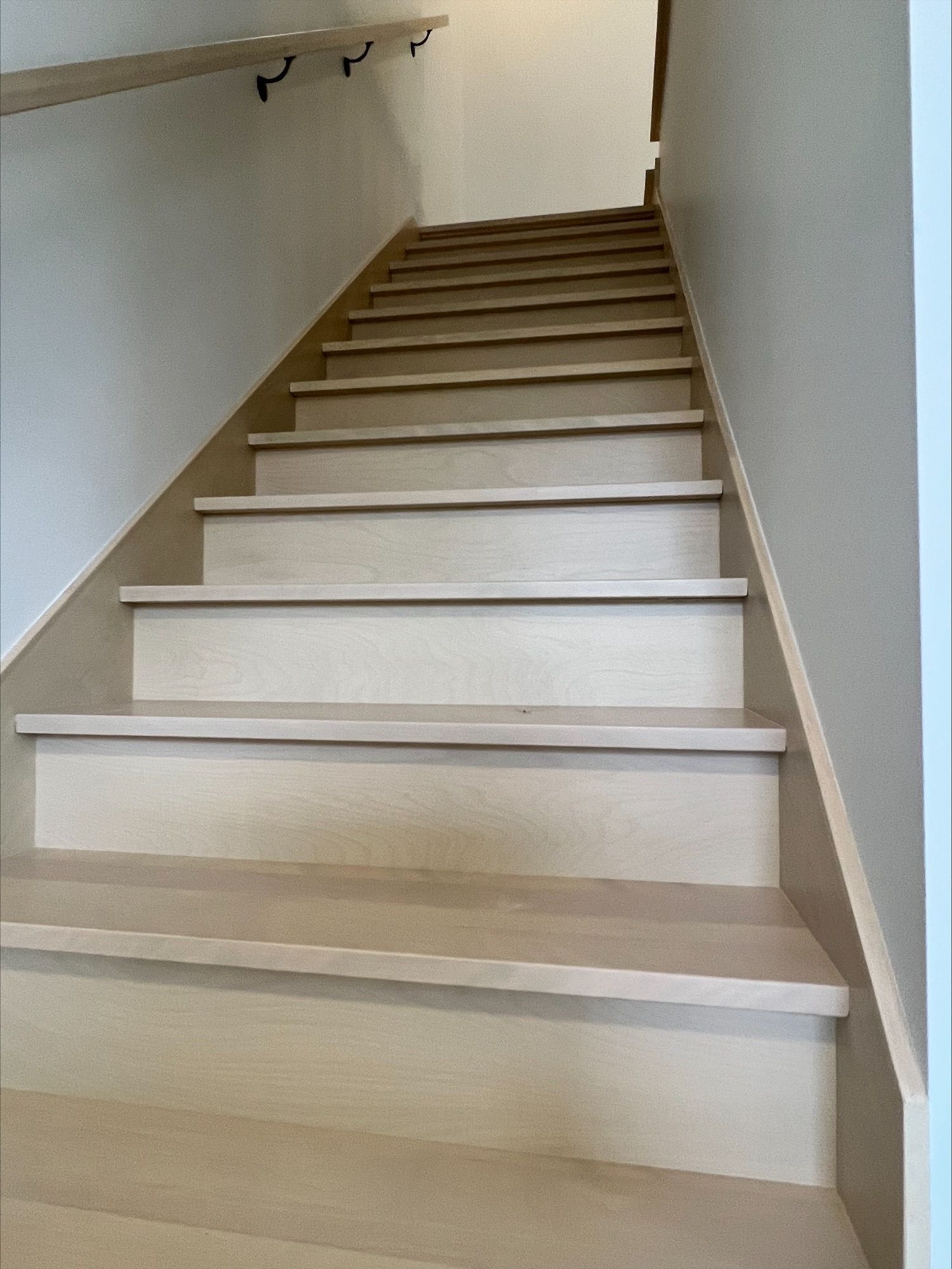 Escalier en bois montant avec marches et murs de couleur claire, une rampe sombre à gauche.