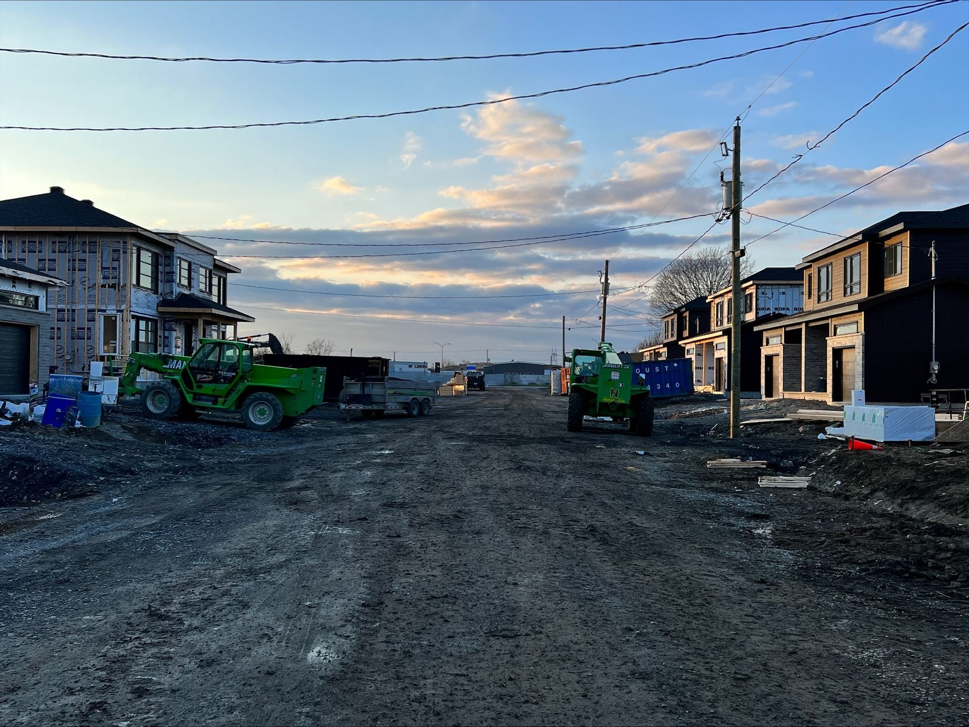 Chantier de construction avec de nouvelles maisons en construction, des machines vertes et un chemin de terre sous un ciel nuageux.