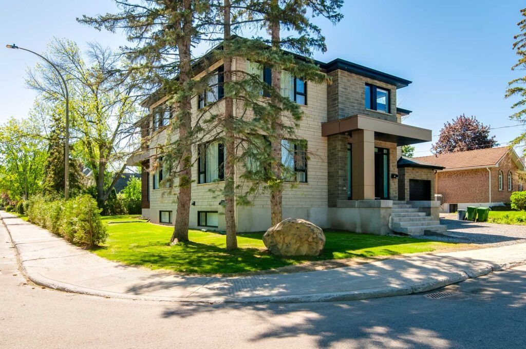 Maison en briques à deux étages avec de l'herbe verte, des arbres et un gros rocher dans la cour avant, vue depuis un coin de rue par une journée ensoleillée.