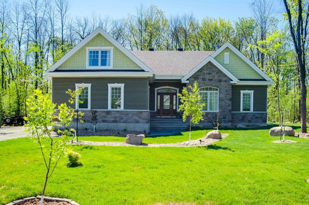 Une maison verte avec des accents de pierre, située sur une pelouse verte et des arbres sous un ciel bleu.