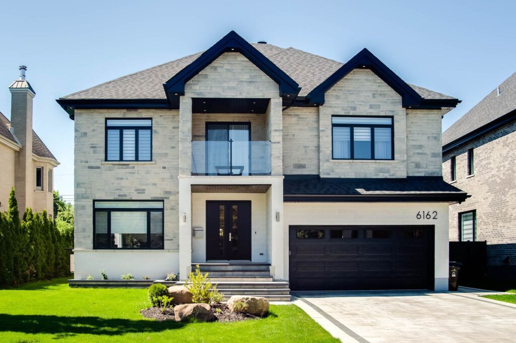 Maison à deux étages avec façade en pierre claire, toit et porte de garage noirs, vue ensoleillée. Pelouse verte et allée pavée.