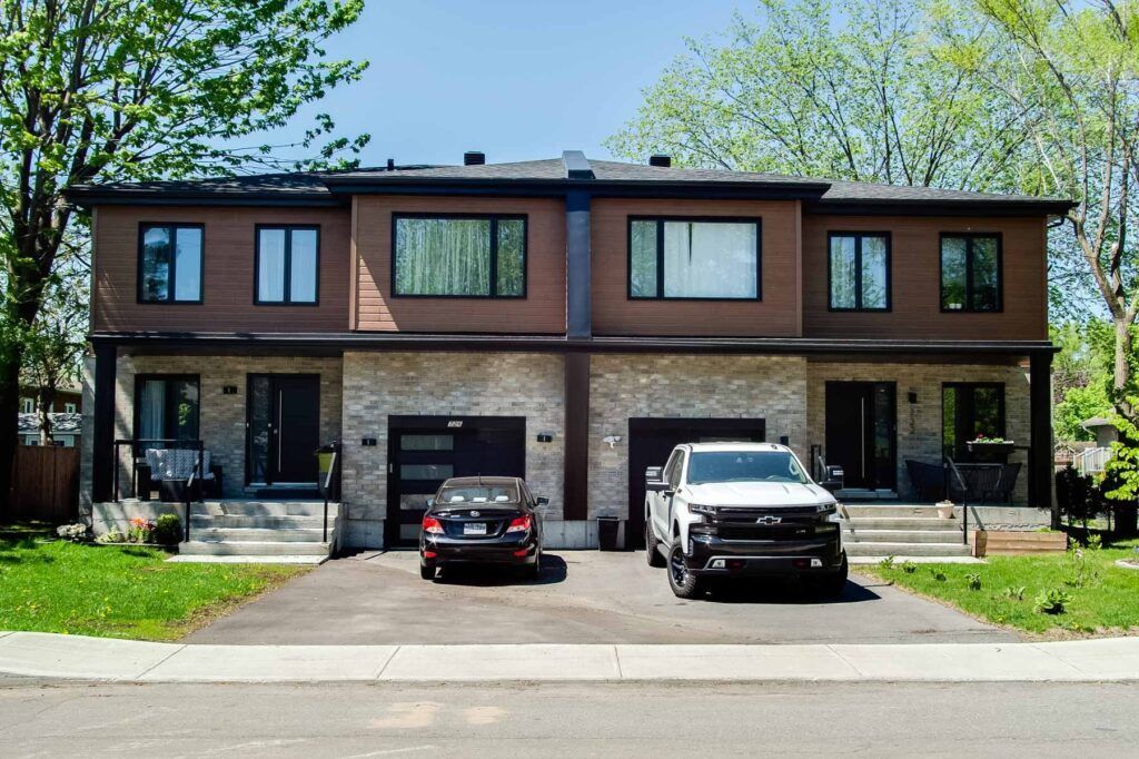Duplex à deux étages avec revêtement brun, touches de pierre et deux portes de garage. Voitures garées dans l'allée.