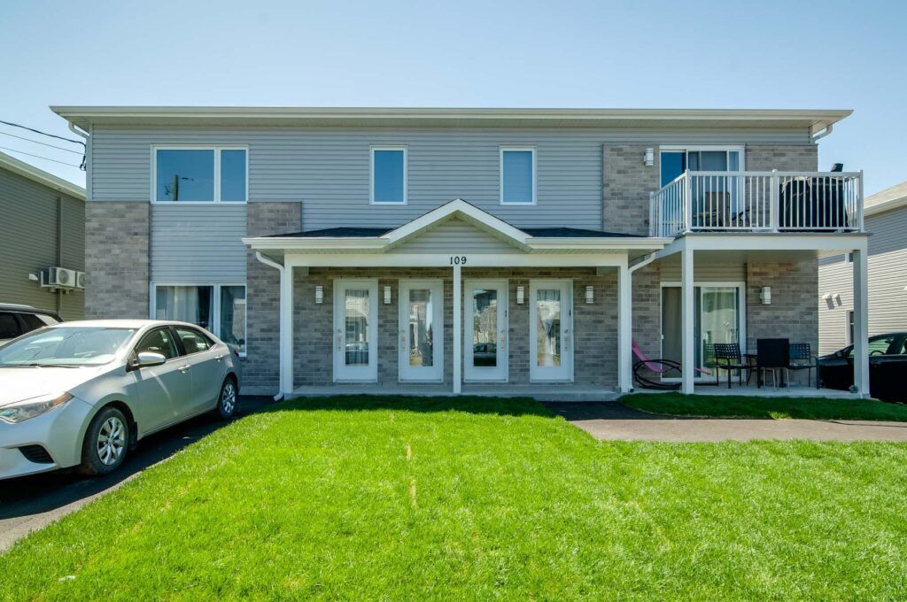 Duplex à deux étages avec revêtement gris clair et touches de brique. Chaque logement dispose d'une porte d'entrée et d'un balcon. Une voiture argentée est garée dans l'allée.