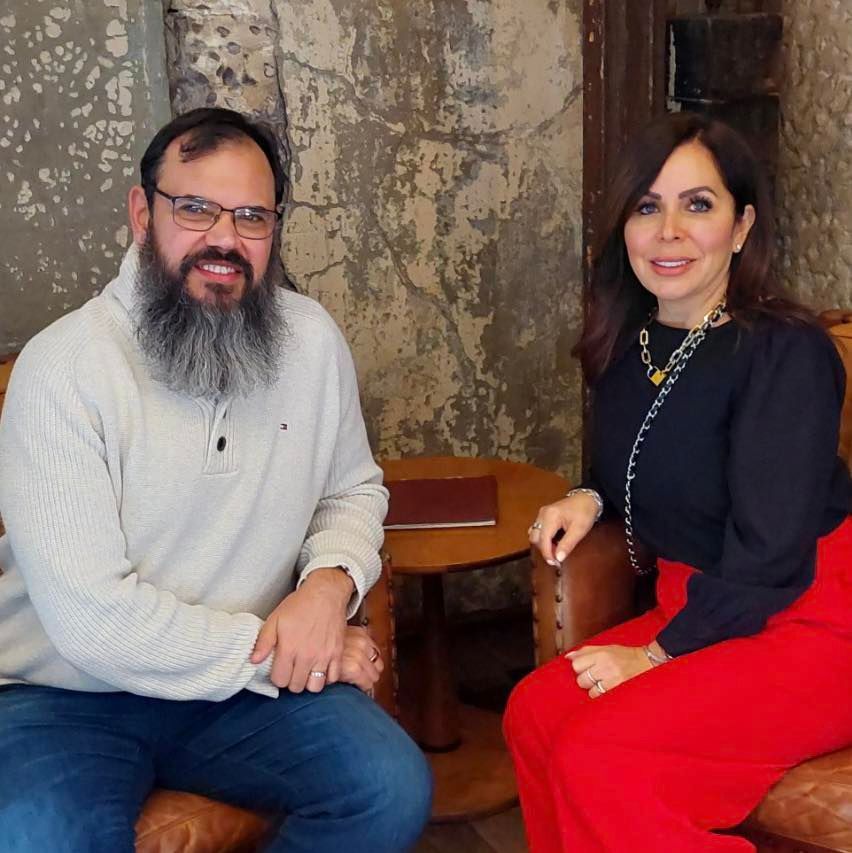 A man with a beard sits next to a woman in red pants