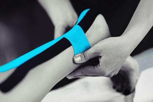 Person applying blue and black kinesiology tape to someone's ankle in a treatment setting.
