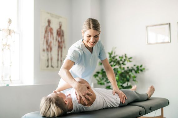 A smiling physical therapist stretches a patient's arm on a treatment table in a bright room.