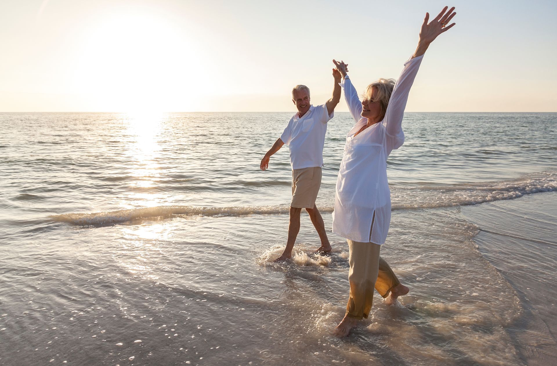 Elderly couple holding hands, dancing on a beach as the sun sets, water splashing around their feet.