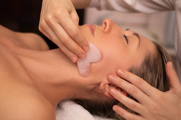 Woman receiving facial Gua Sha treatment with pink tool in a spa setting.