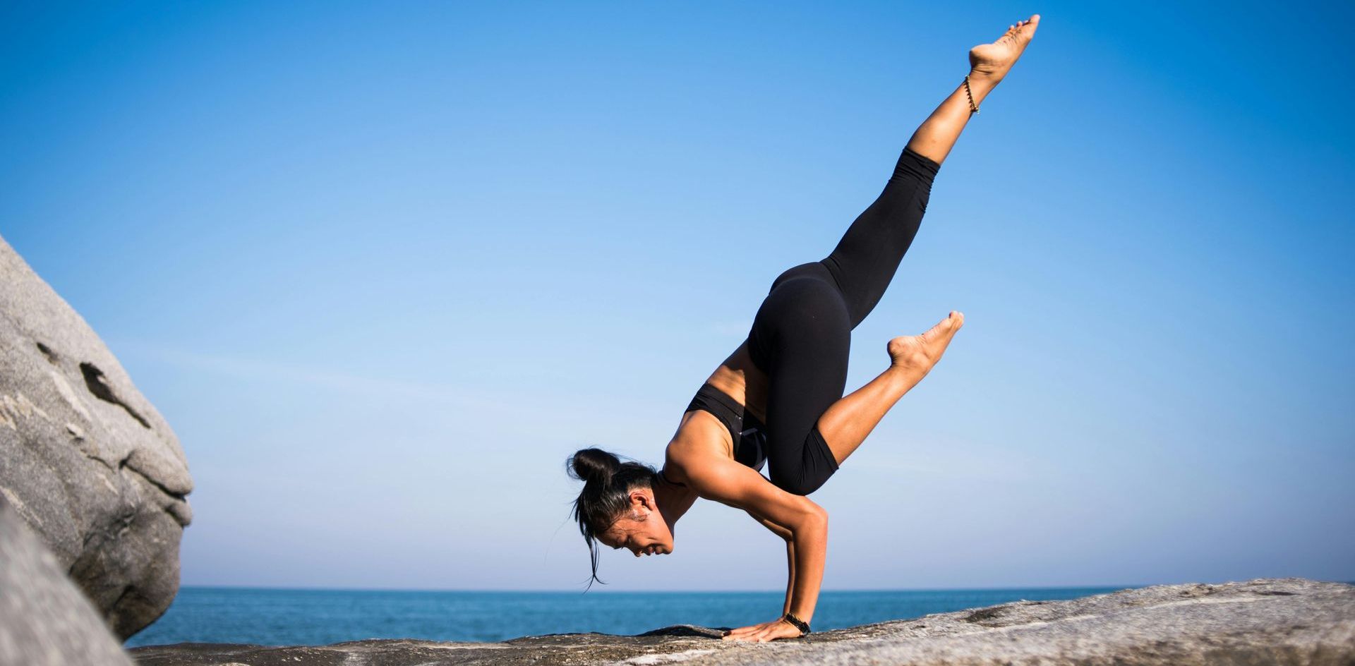 Woman in black activewear doing a handstand on a rock at the beach with a clear blue sky.